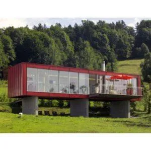 Casa contenedor roja elevada sobre pilares de hormigón, con gran fachada acristalada y bosque detrás