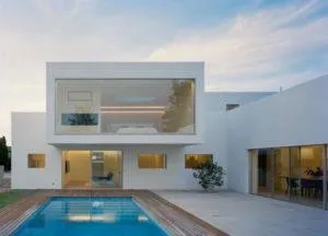 Casa blanca cúbica de dos plantas con gran ventanal superior y piscina rodeada de madera