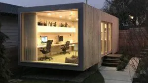 Casa contenedor revestida de madera al anochecer con gran ventanal y despacho iluminado dentro